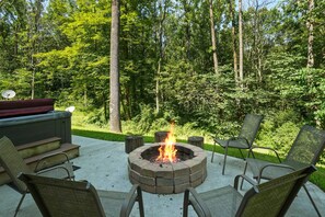 A circular fire pit with chairs around it, set on a concrete patio with a view of a forested area.