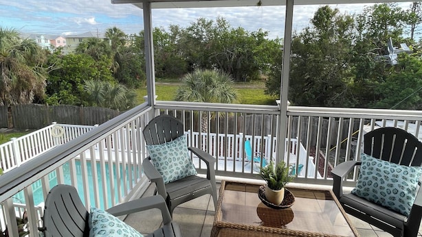 A covered balcony with two chairs and a table, overlooking a swimming pool and greenery. - Gorgeous condo with pool view in walking distance to Siesta Key Beaches (Sarasota)