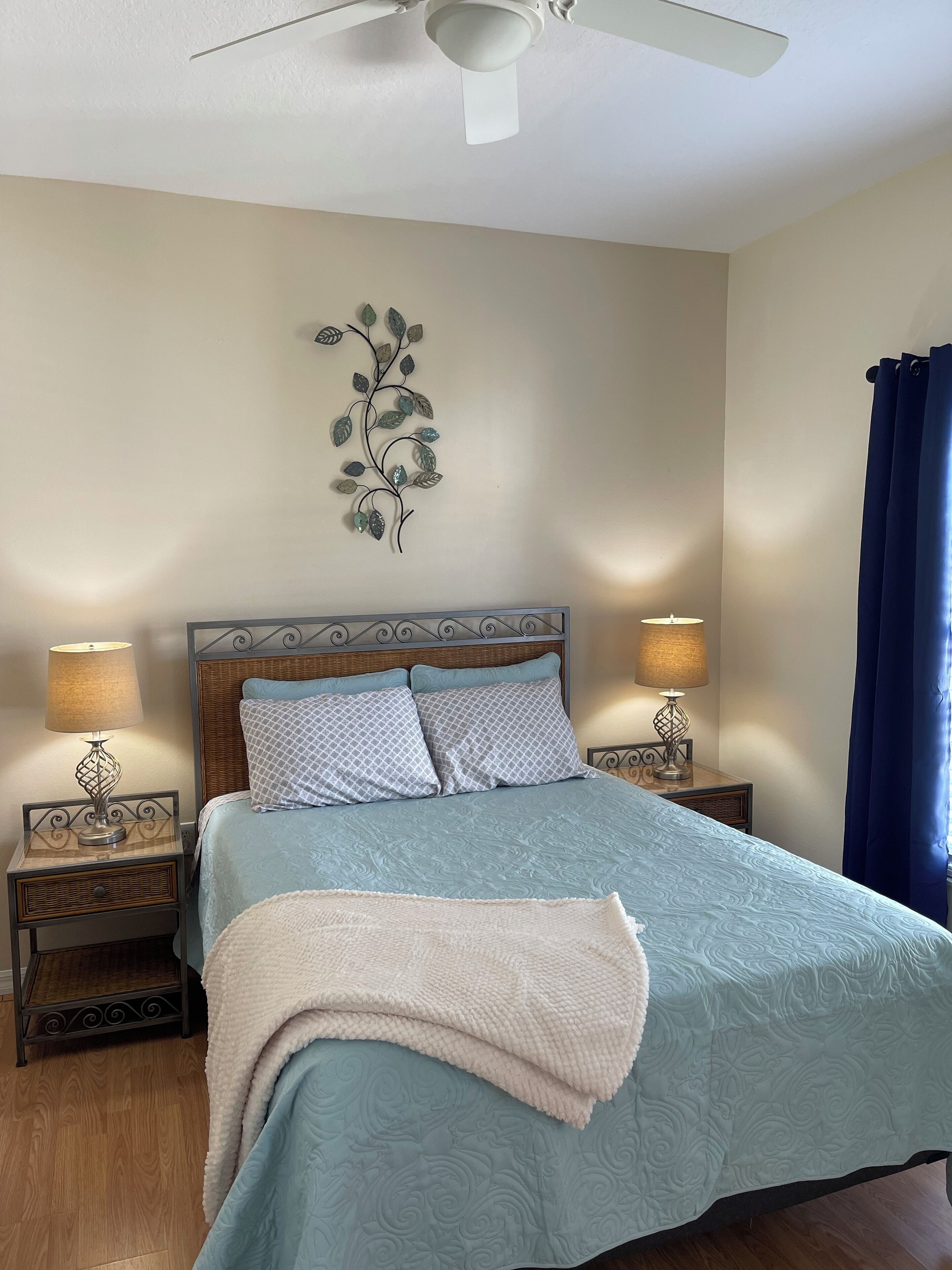 A neatly made bed with a blue bedspread, two patterned pillows, and a decorative wall piece above it. There are two bedside tables with lamps, a ceiling fan, and a window with blue curtains.