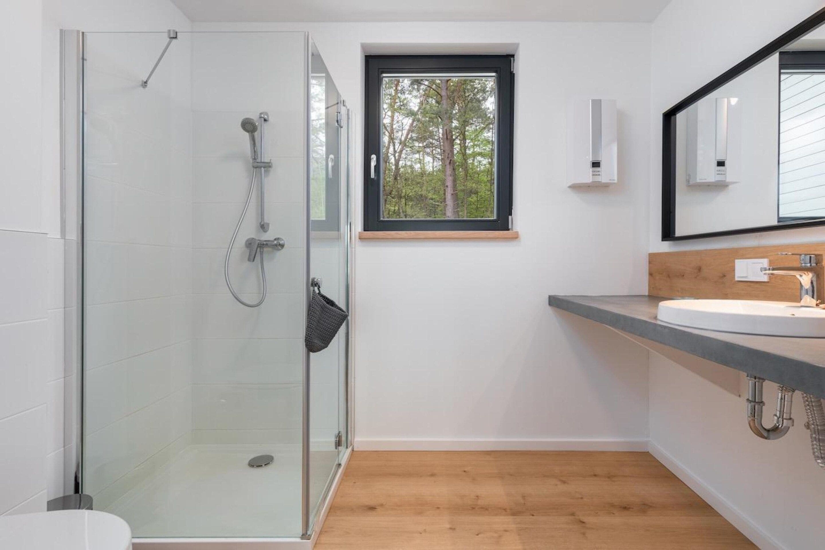 A modern bathroom with a glass shower enclosure, a wooden floor, a white sink, and a window with a view of trees.