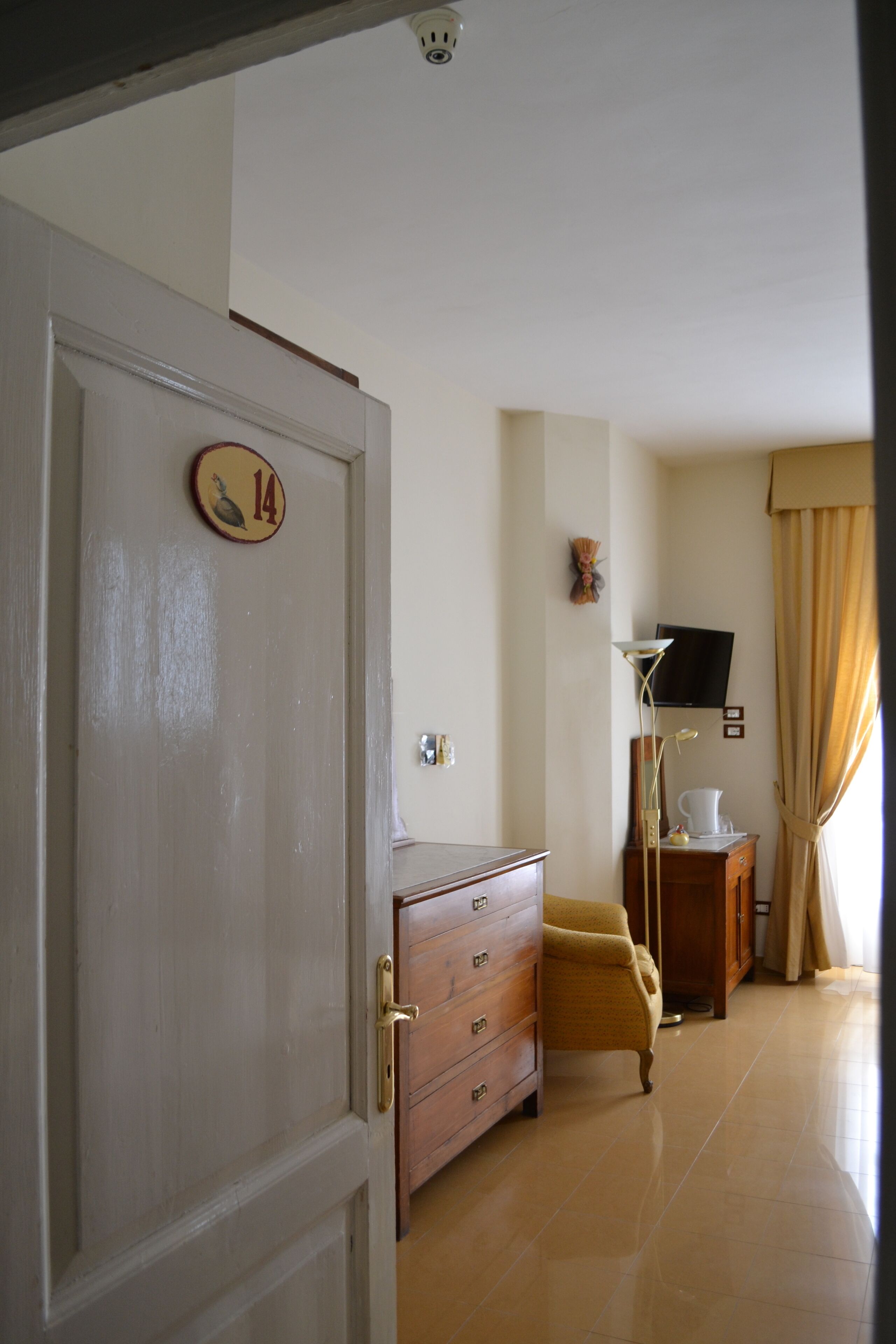 A hotel room with a wooden door marked with the number 14, a wooden dresser, a yellow armchair, a television on a wooden stand, and a window with beige curtains.