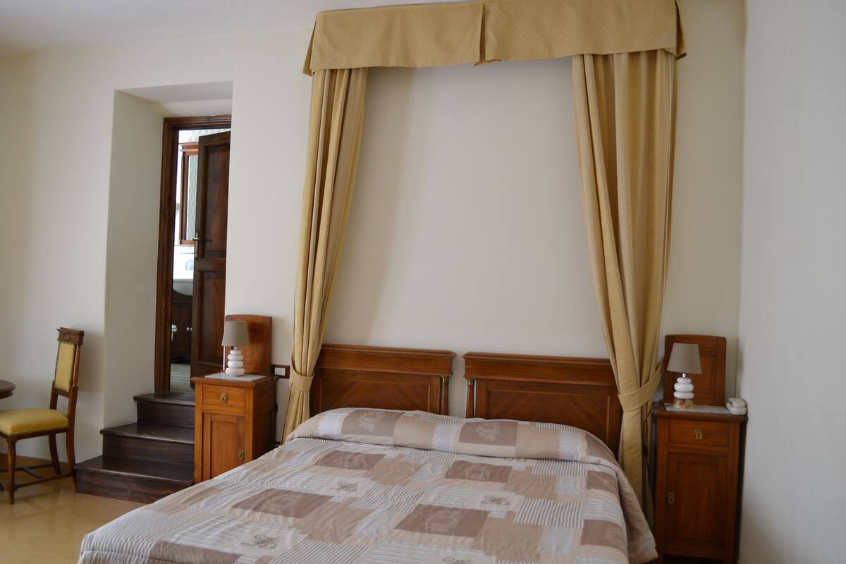 A bedroom with a bed, wooden headboard, bedside tables, a chair, and a staircase leading to another room.