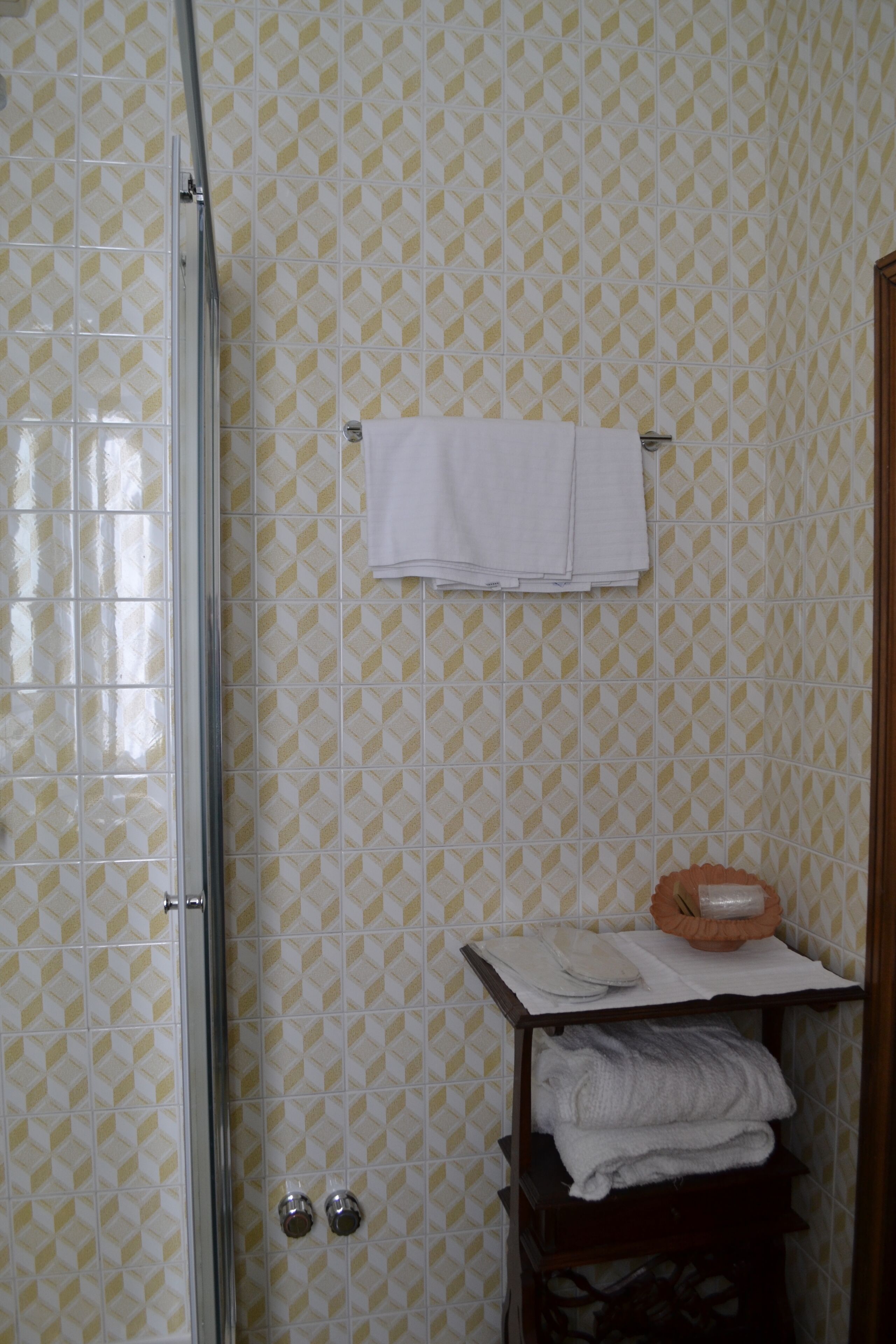 Een badkamer met betegelde vloer, een glazen douchedeur, een handdoekrek met opgevouwen handdoeken, en een plank met opgevouwen handdoeken en een zeepbakje.