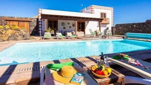 Ein Poolbereich mit Liegestühlen, einem Tisch mit Hut und Buch sowie ein Gebäude mit einer Steinwand und einem Balkon.
