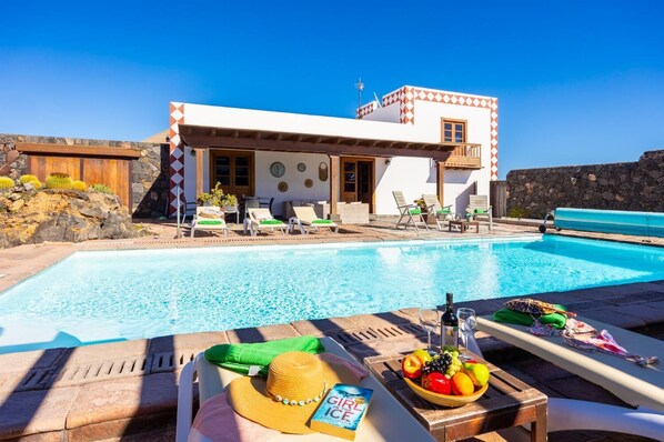 Uma área à beira da piscina com espreguiçadeiras, uma mesa com chapéu e livro, e um edifício com parede de pedra e varanda.