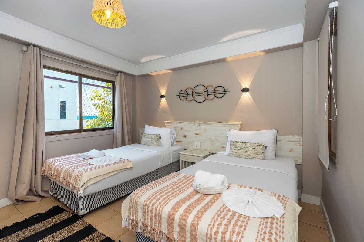 A hotel room with two beds, a large window with curtains, a wall-mounted light fixture, and a hanging shower curtain.
