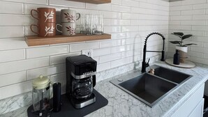 Una cocina con salpicadero de azulejos tipo metro, una cafetera, un lavabo y objetos decorativos en estantes.