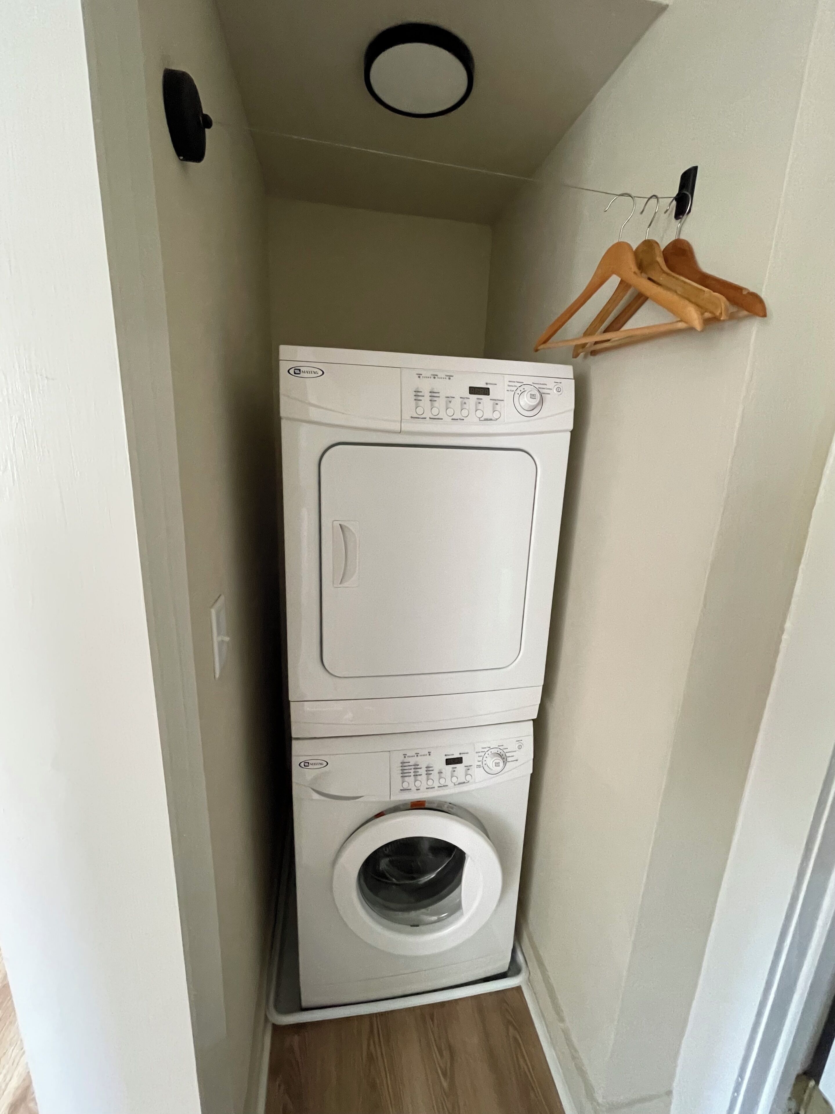 Een compact, wit was- en droogcombitoestel in een kleine wasruimte, met een houten hangerrek erboven.