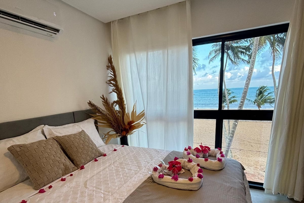 A hotel room with a bed, a large window with a view of the beach and palm trees, and a potted plant on the windowsill.