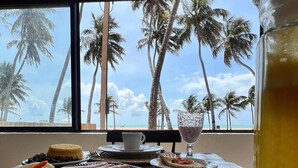 Un bufé de desayuno con diversas opciones de comida y vista a palmeras y la playa a través de amplias ventanas.