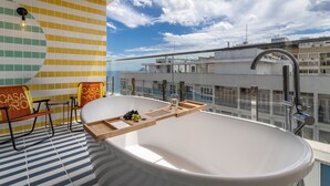 A modern bathroom with a freestanding bathtub, a wooden tray with fruits and a glass, and a view of the cityscape. - Sea View and Balcony with Bathtub | Q5 (Rio de Janeiro)