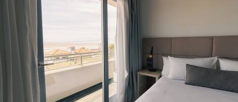 A hotel room with a large window, a bed with white and grey bedding, a bedside table with a lamp, and a view of buildings outside.