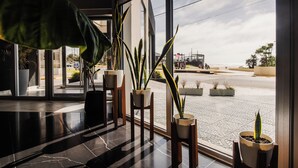 A modern lobby with large windows, potted plants, and a view of the street and beach.