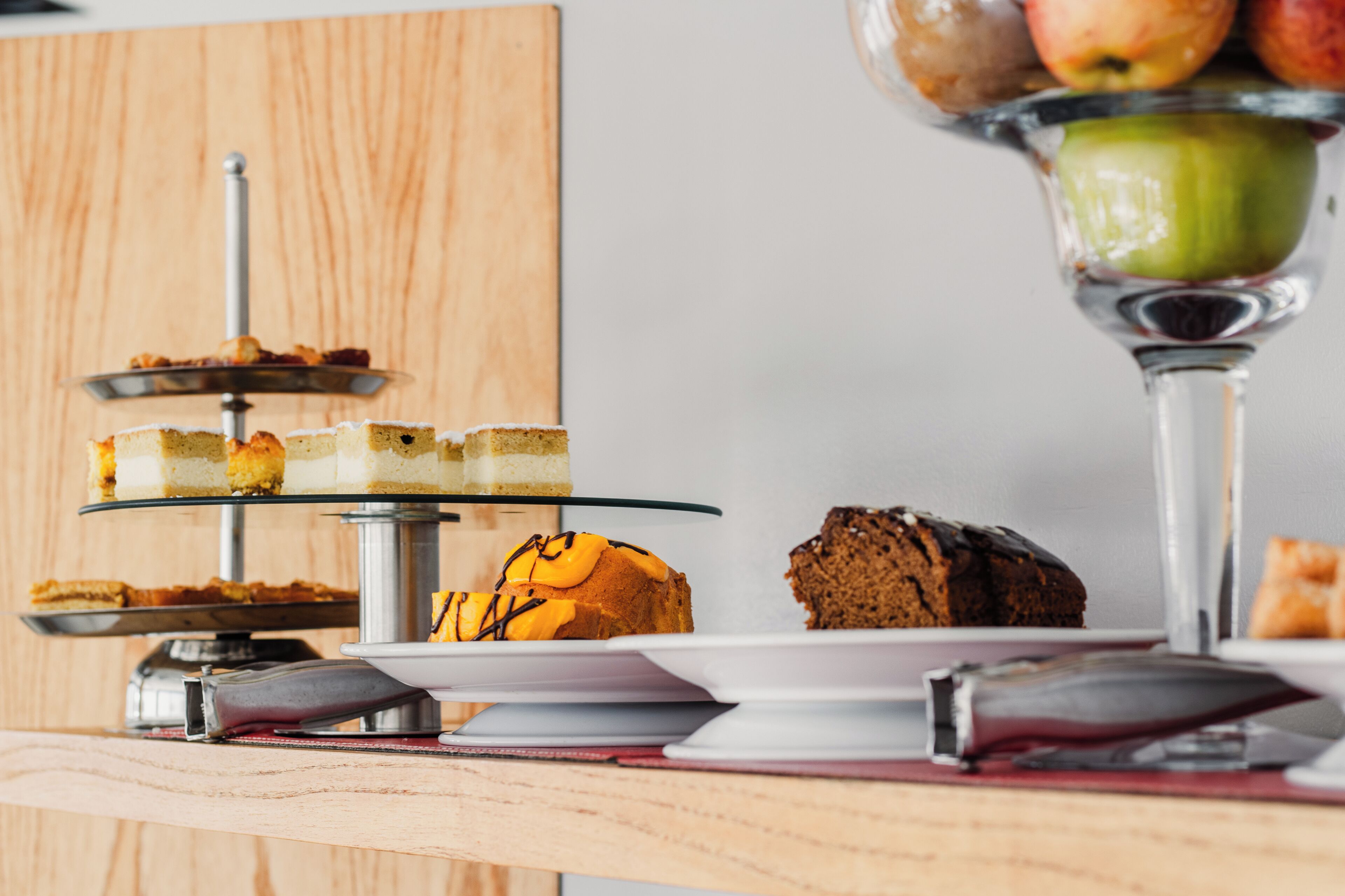 Un présentoir à gâteaux à trois niveaux, garni de divers gâteaux et pâtisseries, un saladier en verre rempli de pommes et un livre posé sur la table.