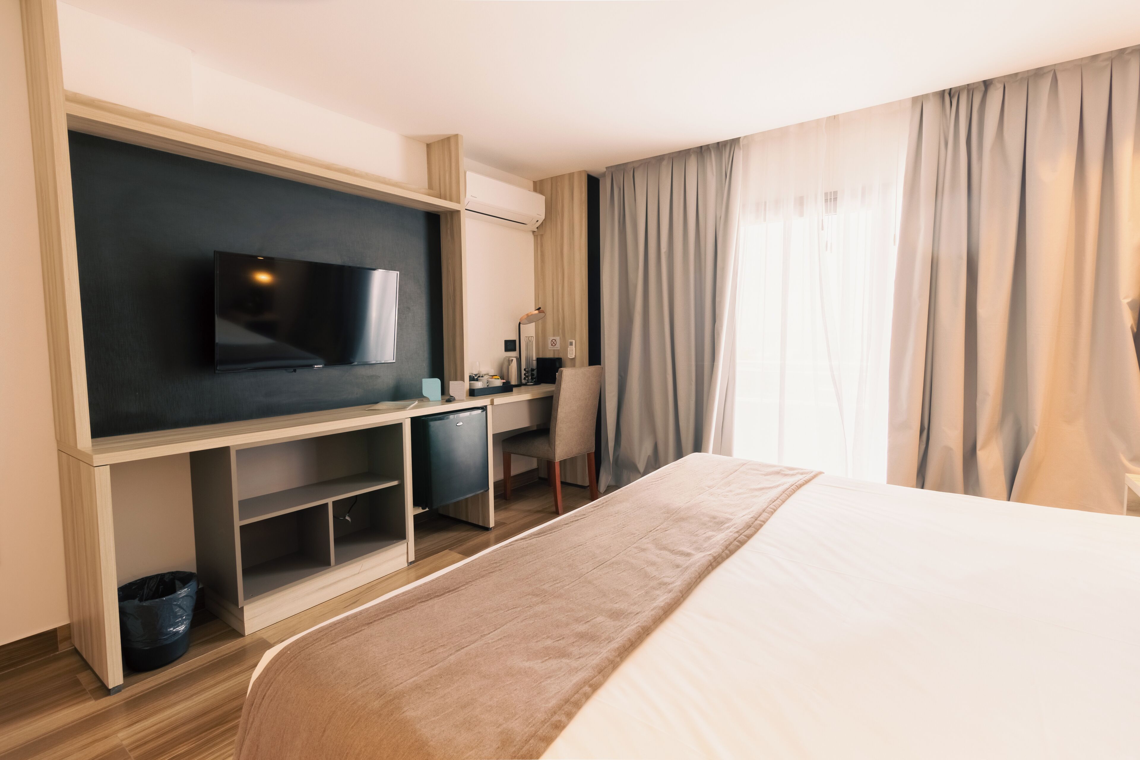 Ein Hotelzimmer mit einem Bett, einem Schreibtisch mit Computer, einem Fernseher und einem Fenster mit Vorhängen.