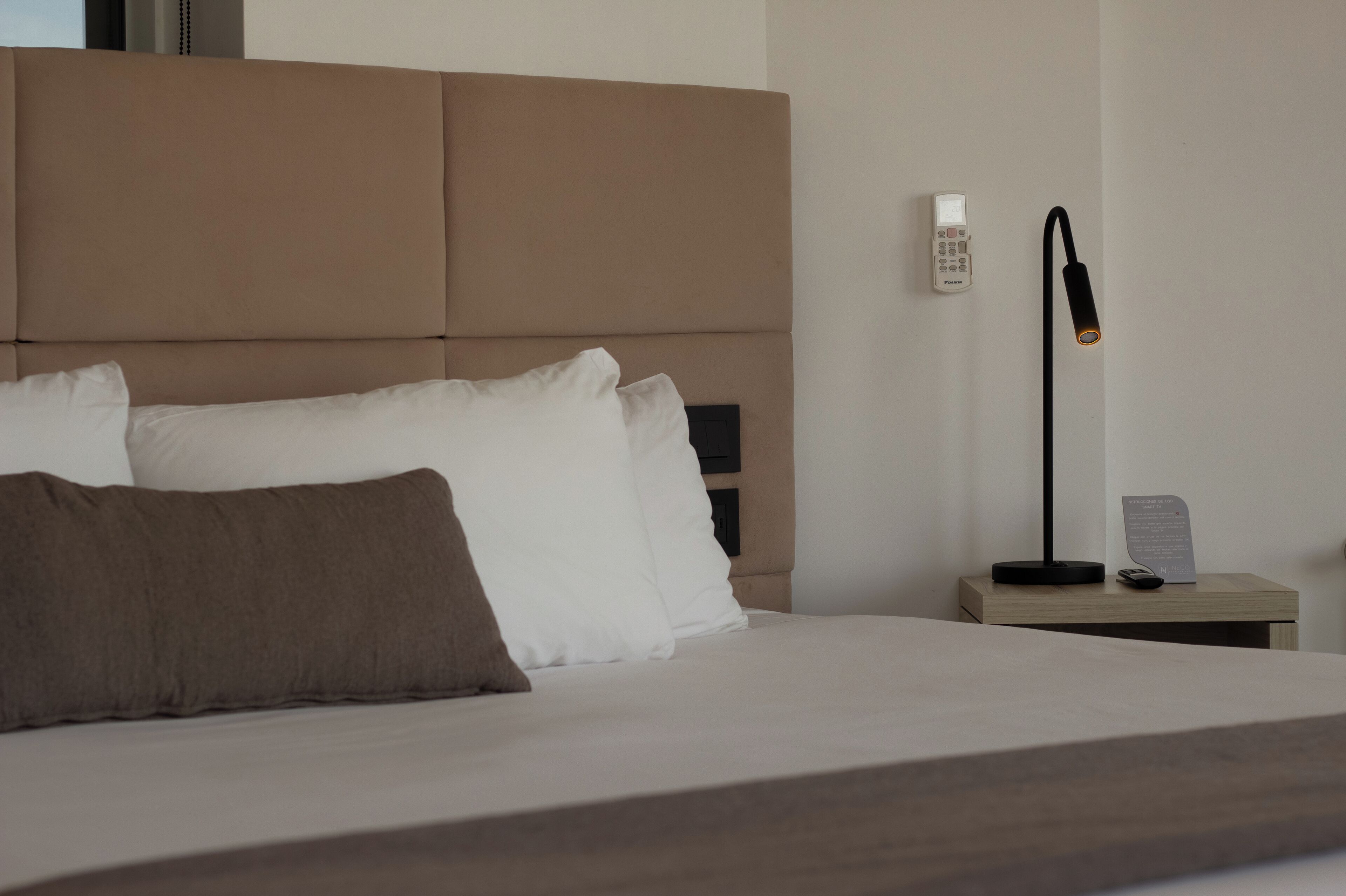 Una cama bien hecha con almohadas blancas y marrones, una mesita de noche con una lámpara y un pequeño cartel en la pared.