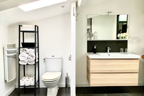 Une salle de bain moderne avec des toilettes, une étagère noire avec des serviettes, un miroir au-dessus du lavabo et un sol carrelé foncé.