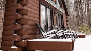 Eine Blockhütte mit Veranda und zwei Fenstern, umgeben von Schnee und Bäumen.
