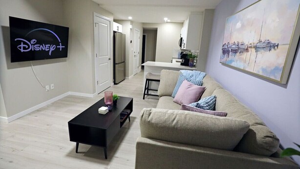 A modern living room with a flat-screen TV displaying Disney+, a black coffee table, a beige sofa, and a painting of boats on the wall. - Edgemont Escape Suite|Long Stays|WEM|Disney+ (Edmonton)