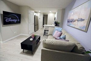 A modern living room with a flat-screen TV displaying Disney+, a black coffee table, a beige sofa, and a painting of boats on the wall. - Edgemont Escape Suite|Long Stays|WEM|Disney+ (Edmonton)
