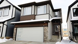 A two-story house with a garage, stone foundation, and a snow-covered driveway. - Edgemont Escape Suite|Long Stays|WEM|Disney+ (Edmonton)