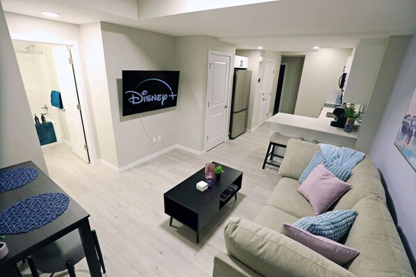 A modern living room with a flat-screen TV mounted on the wall, a black coffee table, a beige sofa with patterned cushions, and a kitchen area in the background. - Edgemont Escape Suite|Long Stays|WEM|Disney+ (Edmonton)