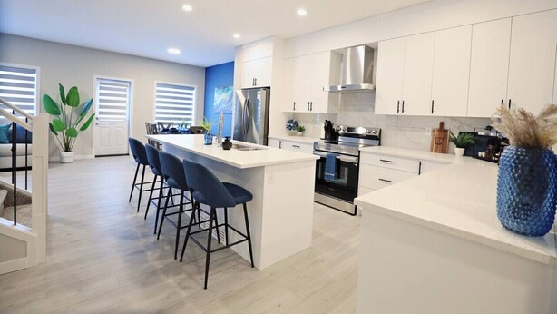 A modern kitchen with white cabinets, a central island with bar stools, stainless steel appliances, and a blue vase on the counter. - Edgemont Escape |A/C|Groups|River Cree|WEM|Disney+ (Edmonton)