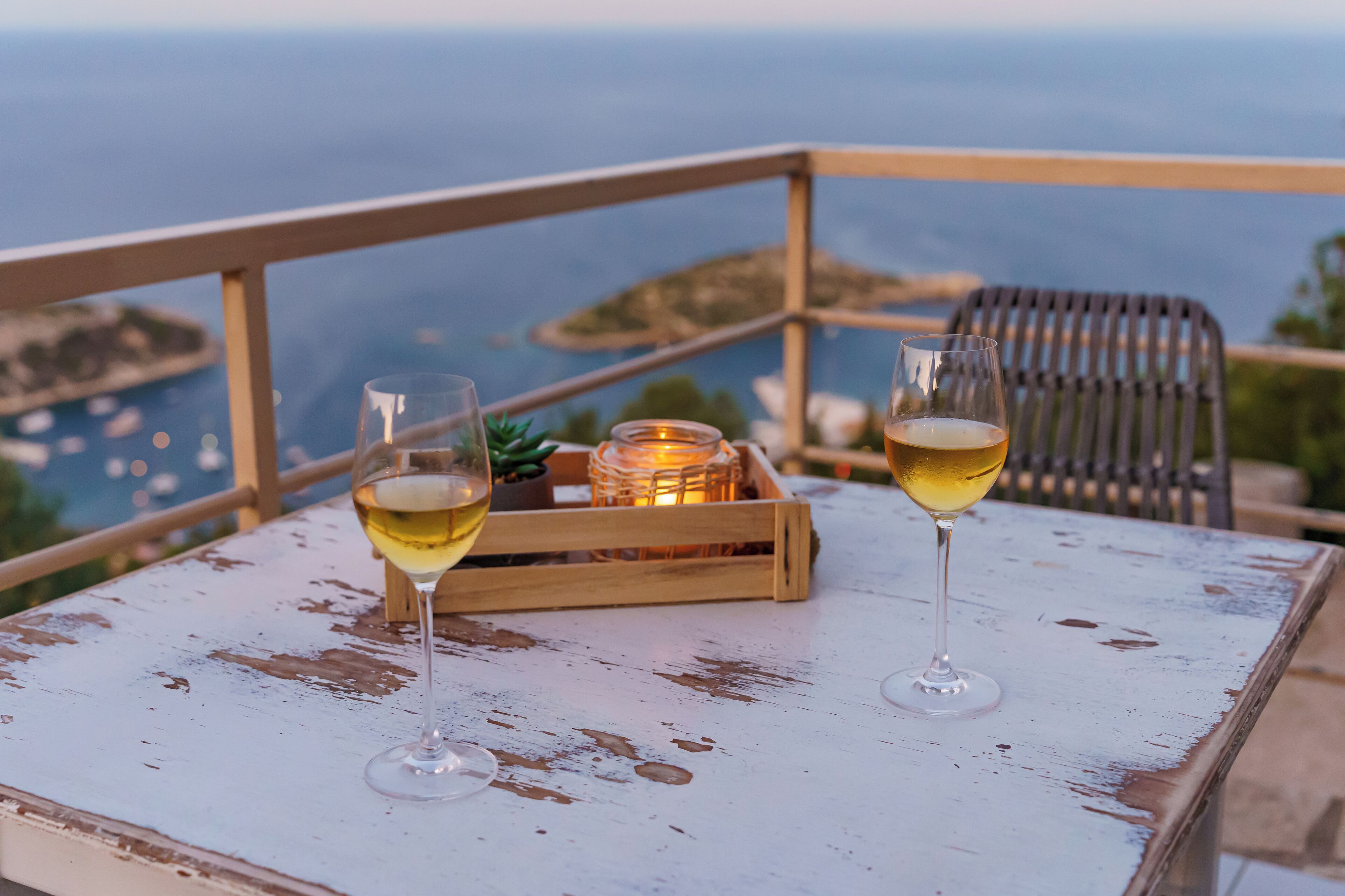Zwei Gläser Weißwein auf einem weiß gestrichenen Tisch mit Blick auf das Meer und eine entfernte Insel.
