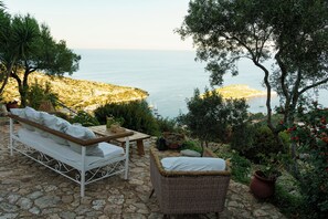 Une terrasse en pierre avec un canapé en métal blanc et un fauteuil en osier, offrant une vue sur un paysage côtier avec des bateaux et une île au loin.