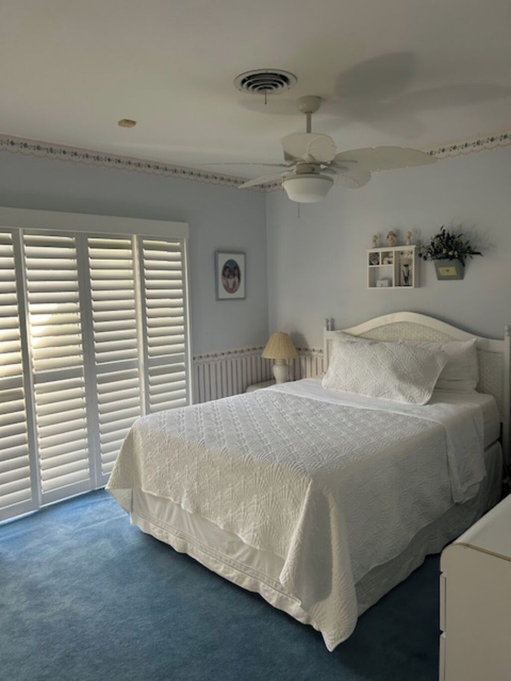 Ein Schlafzimmer mit Bett, weißen Wänden, Deckenventilator und Fenster mit Jalousien.