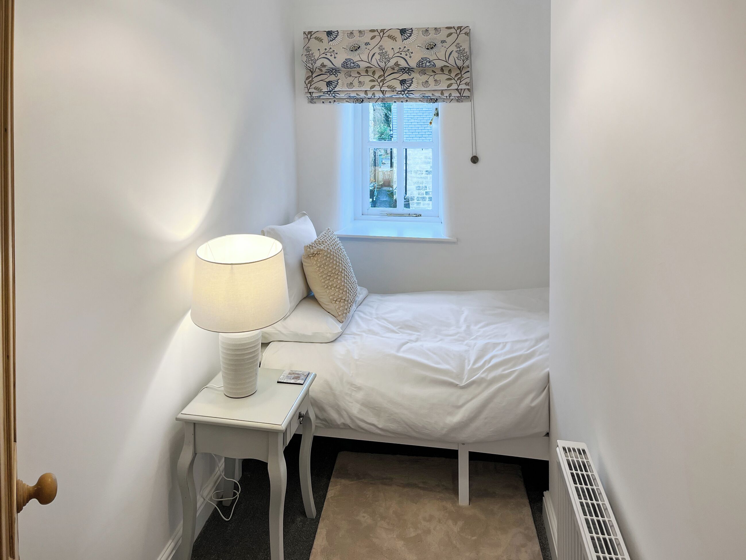 Une petite chambre à coucher blanche, avec un lit simple, une table de chevet avec une lampe, une fenêtre agrémentée d’une lambrequin à motifs floraux et un radiateur.