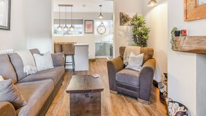 A modern living room with a sofa, armchairs, and a coffee table. A kitchen is visible in the background with pendant lights and a clock. - Hook Cottage - UK48361 (Loftus, near Staithes)