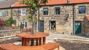 A stone building with a red-tiled roof, a circular wooden table with benches, and a small tree in the center. - Hook Cottage - UK48361 (Loftus, near Staithes)
