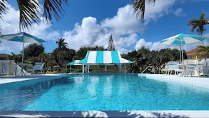 A clear blue pool with white and blue striped umbrellas, surrounded by palm trees and a clear sky. - Condo - 2 beds with WiFi, AC, Pool and Oceanfront in charming Frigate Bay. (Frigate Bay)