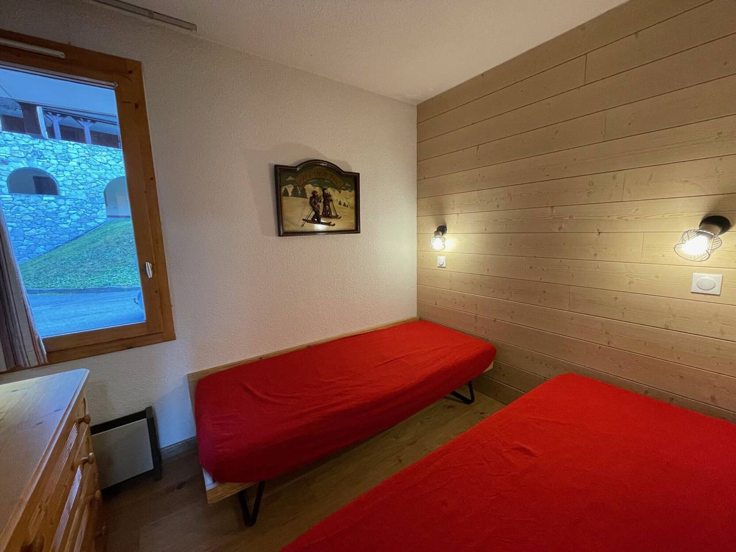 A room with two single beds, a wooden wall, a framed picture, a window, and a wall-mounted light fixture.