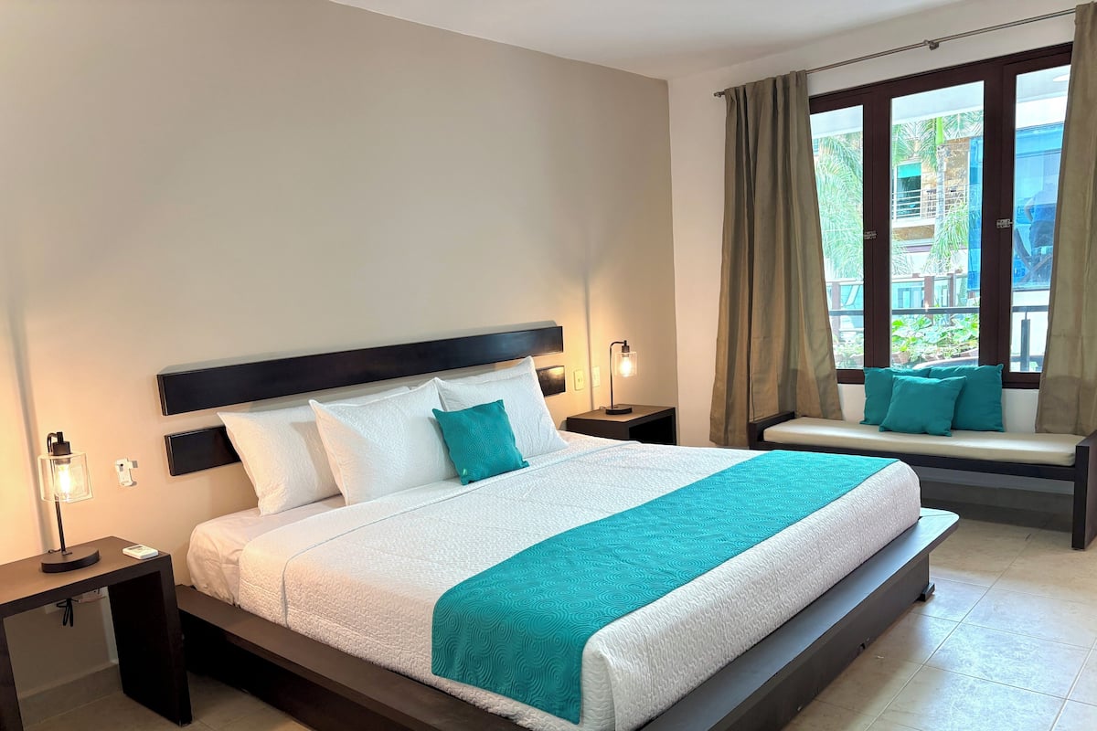 A neatly arranged hotel room with a large bed, a bedside table, a reading lamp, and a window with curtains.