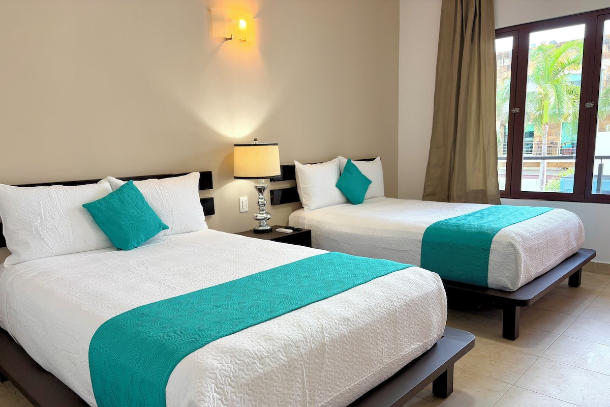 A hotel room with two beds, each with white bedding and teal accents, a nightstand with a lamp, and a window with a view of palm trees.