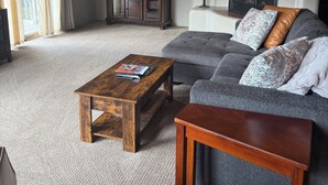 Una sala de estar con techo de madera, un sofá gris con cojines estampados, una mesa de centro de madera y un mueble con espejo y objetos decorativos.