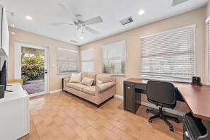 A room with a beige sofa, a desk with a chair, a ceiling fan, and large windows with blinds.