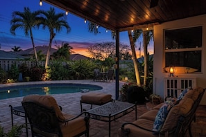 Un espace aménagé au bord de la piscine, avec un patio couvert, du mobilier d’extérieur et une vue sur le coucher de soleil.