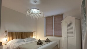 A bedroom with a bed, bedside lamps, a wooden headboard, a window with blinds, a wardrobe, and a small wooden stool.