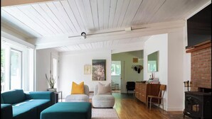 A modern living room with a teal sofa, a white armchair, and a wooden floor. There is a fireplace on the right and a desk with a chair in the background. - ​Bright and Spacious Secluded Forest Home with Sprawling Yard and Private Brook (Cornwall)