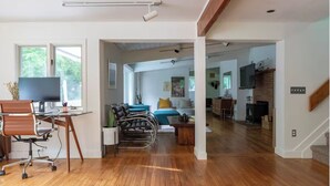 A spacious living area with a wooden floor, a desk with a computer, a sofa, and a dining area with a brick wall and a fireplace. - ​Bright and Spacious Secluded Forest Home with Sprawling Yard and Private Brook (Cornwall)