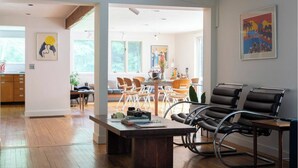 A modern living room with a wooden floor, a dining area with chairs, and a kitchen in the background. - ​Bright and Spacious Secluded Forest Home with Sprawling Yard and Private Brook (Cornwall)