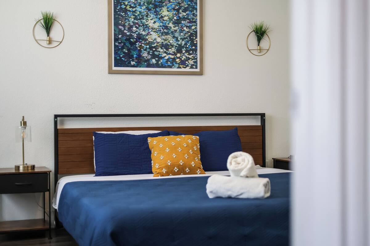 Un lit bien fait, agrémenté d’oreillers bleus et jaunes, un tableau abstrait encadré au-dessus, et deux petites plantes en pot sur les tables de chevet.