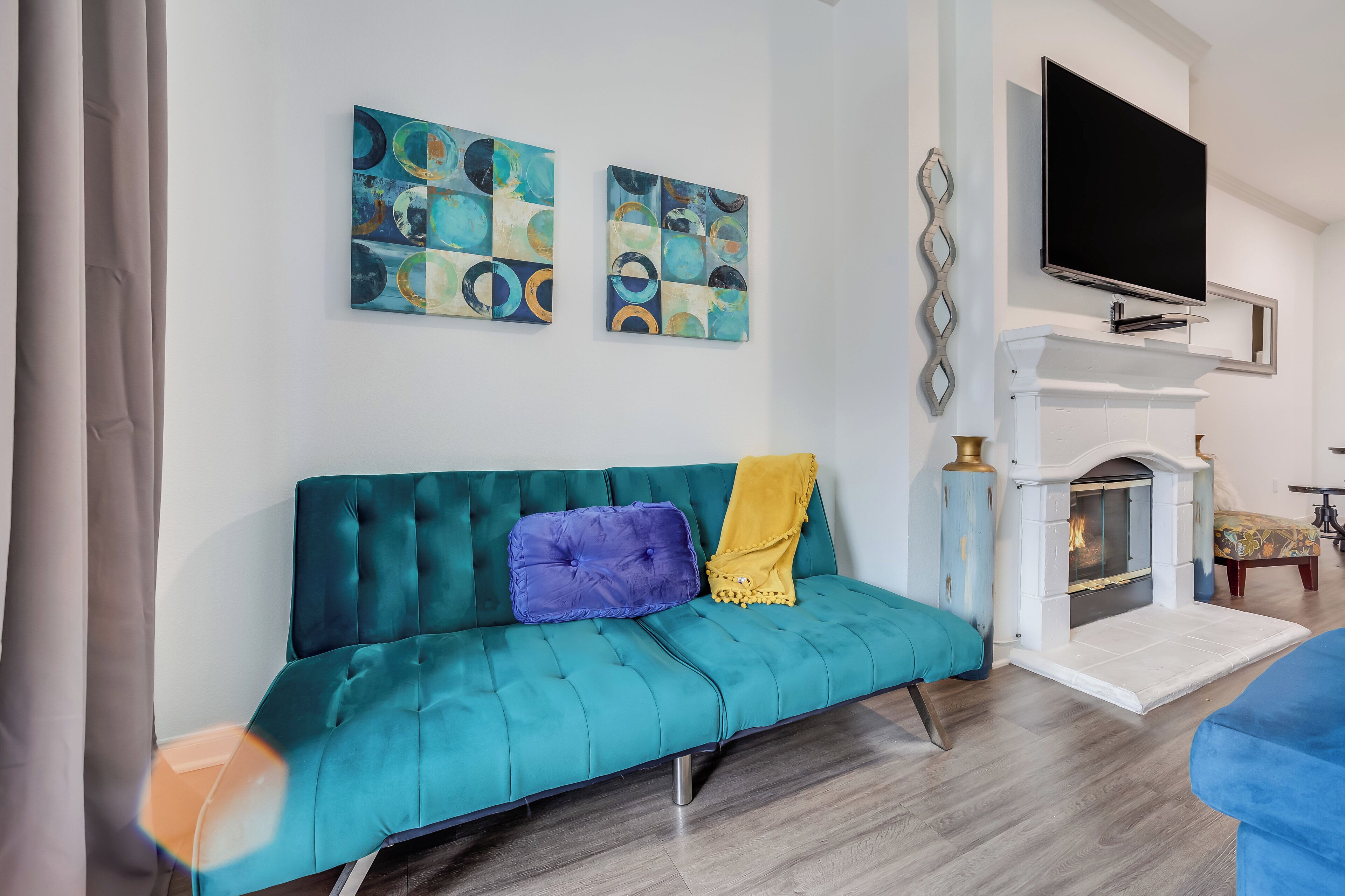 Un canapé de couleur turquoise agrémenté d’un coussin aux motifs violets et jaunes, un téléviseur à écran plat fixé au-dessus d’une cheminée, et des œuvres d’art abstraites accrochées au mur.