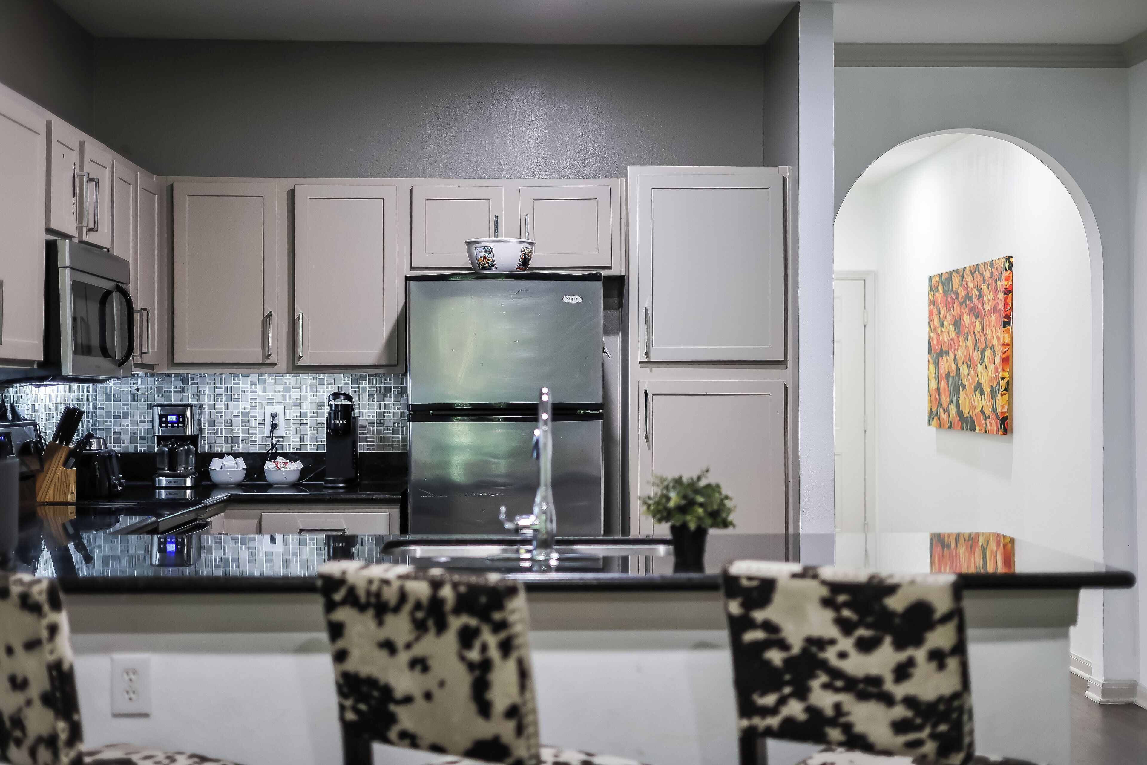 A modern kitchen with a stainless steel refrigerator, a microwave, and a coffee maker. There is a dining area with a glass table and patterned chairs.
