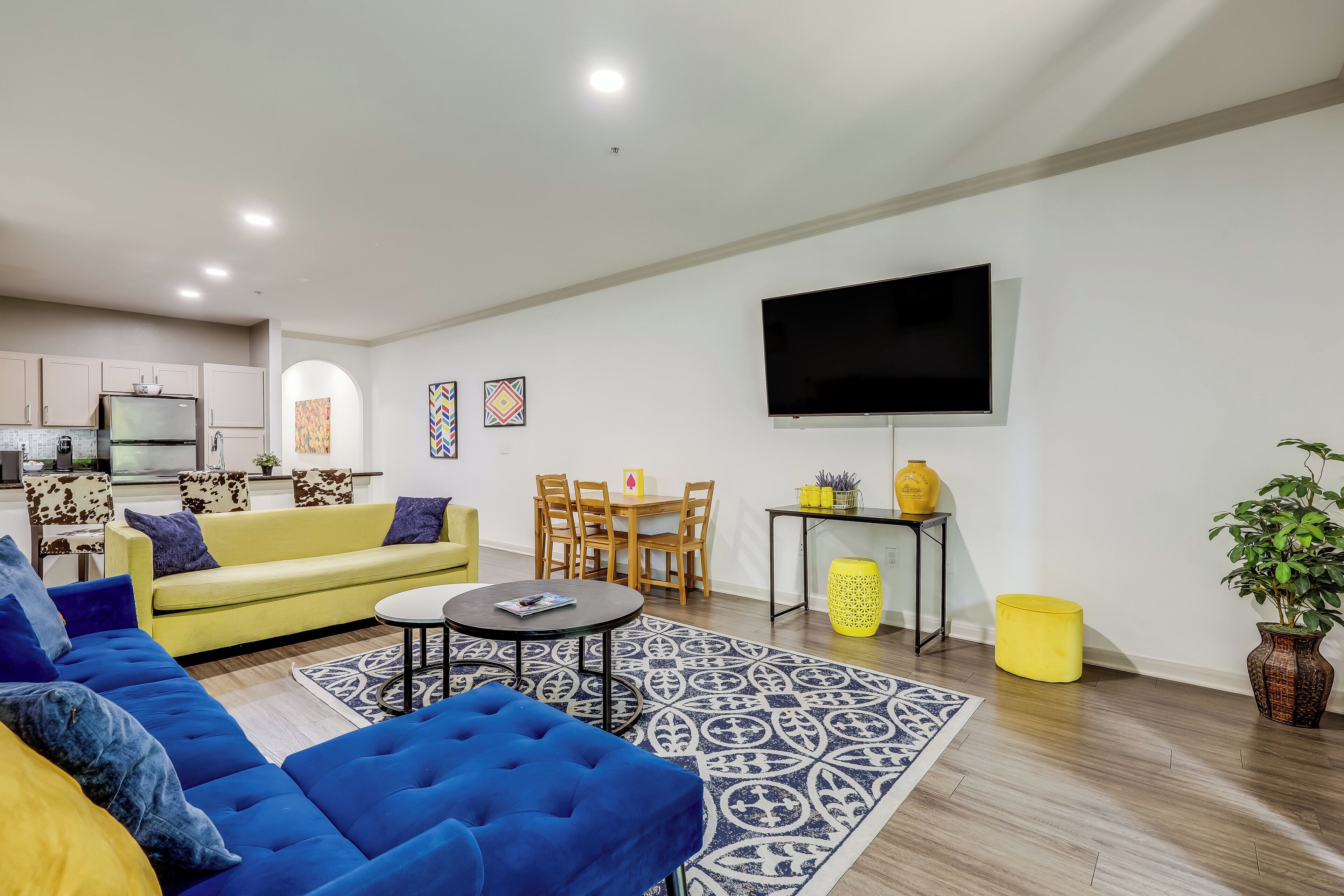 A modern living room with a yellow sofa, blue sofa, and a TV mounted on the wall.