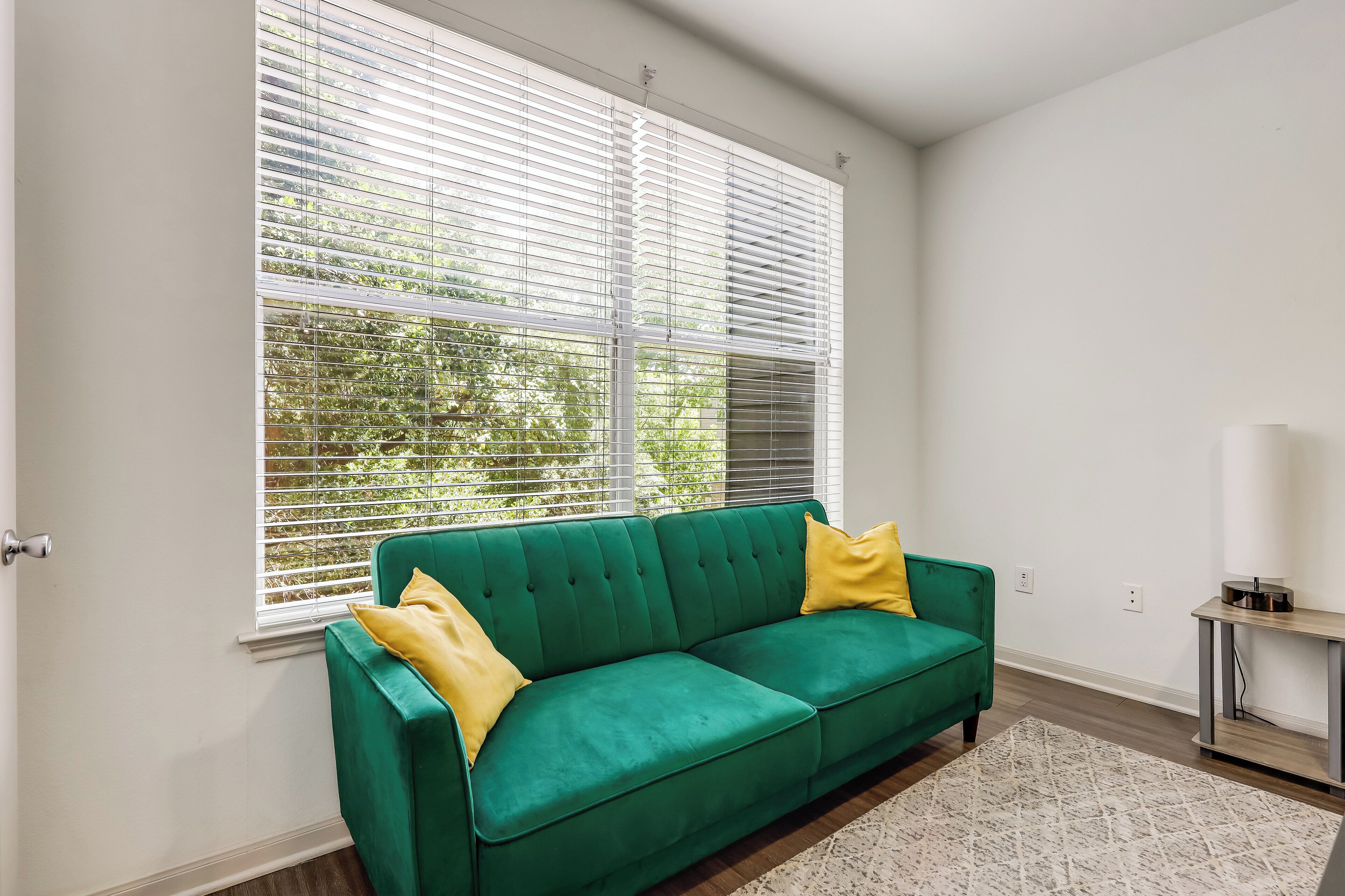 A green sofa with yellow cushions, a white wall, and a window with blinds.