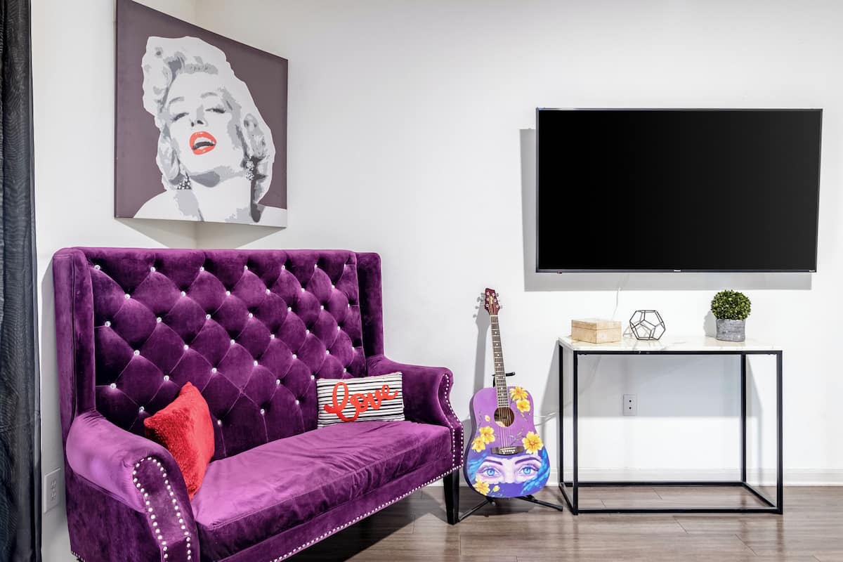 Une pièce avec un canapé en velours violet, une guitare, un téléviseur et une petite table ornée d’objets décoratifs.